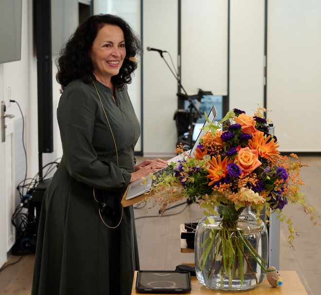 Frau hält eine Rede vor Publikum, neben einem bunten Blumenstrauss.
