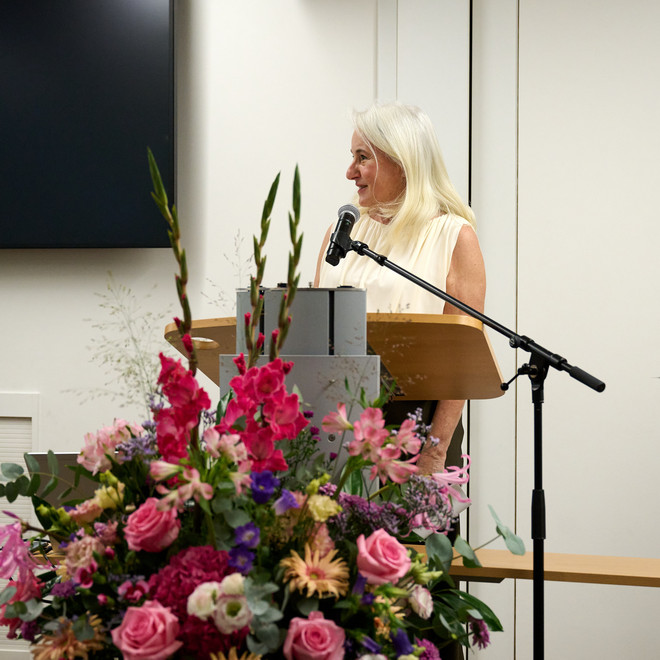 Frau hält eine Rede am Mikrofon hinter einem Rednerpult mit Blumenschmuck.