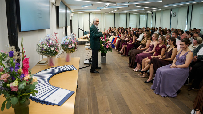 Redner spricht vor den Gästen einer Diplomfeier, während die Diplome bereitliegen.