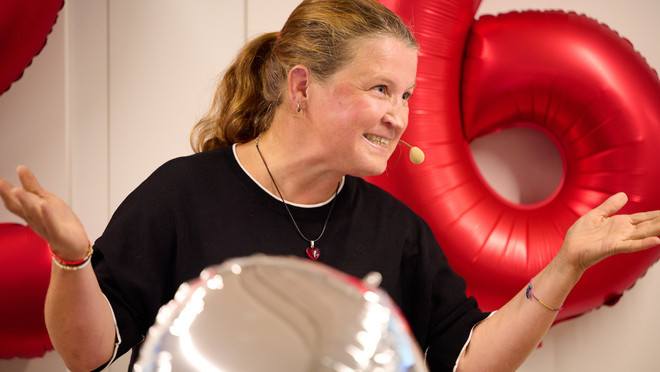 Rednerin mit Mikrofon vor roten Zahlenballons und silbernen Herzballons.