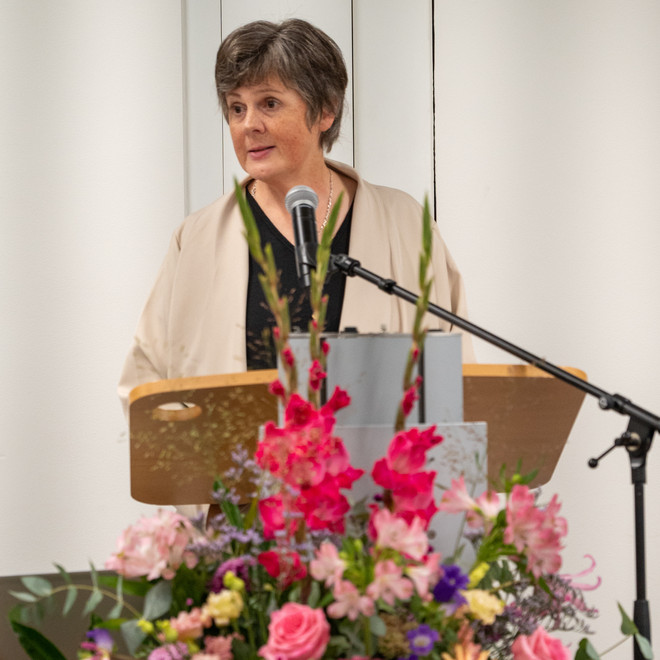 Frau hält eine Rede am Rednerpult, geschmückt mit einem bunten Blumenarrangement.