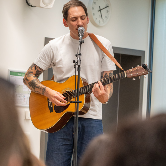 Musiker singt ins Mikrofon und begleitet sich auf einer Akustikgitarre.
