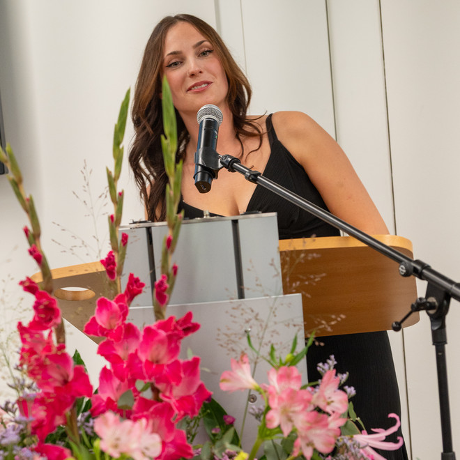 Junge Frau im schwarzen Kleid hält eine Rede hinter einem blumengeschmückten Rednerpult.